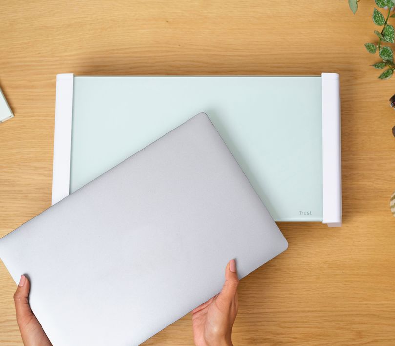 Una persona está colocando una laptop plateada sobre un soporte para monitor de cristal templado Trust Monta de color blanco, sobre un escritorio de madera