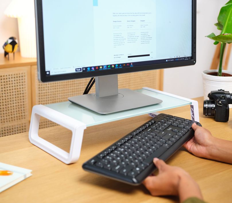 Una persona guardando un teclado debajo de un Soporte para monitor de cristal templado Trust Monta de color blanco, en un escritorio de madera. Hay una planta y una cámara fotográfica al lado del monitor.