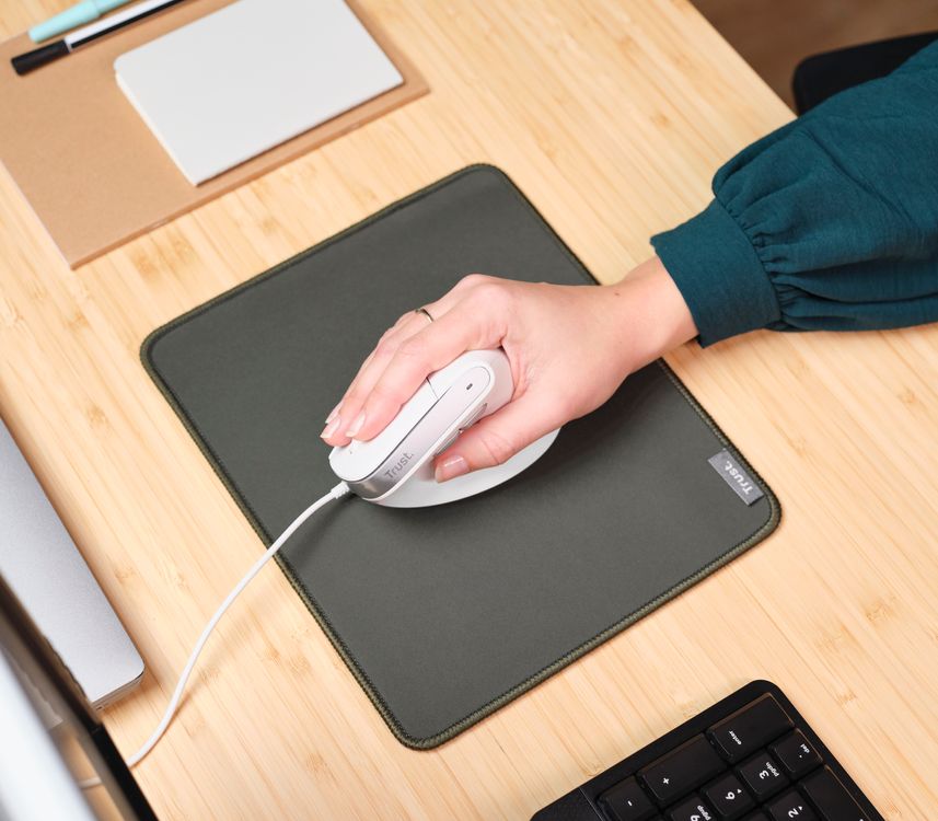 Una mano usando un mouse ergonómico Trust Bayo II de color blanco con cable sobre una alfombrilla verde en un escritorio de madera clara. En la esquina superior izquierda se ven algunos artículos de papelería, como un cuaderno y bolígrafos y en la esquina inferior derecha, un teclado negro.