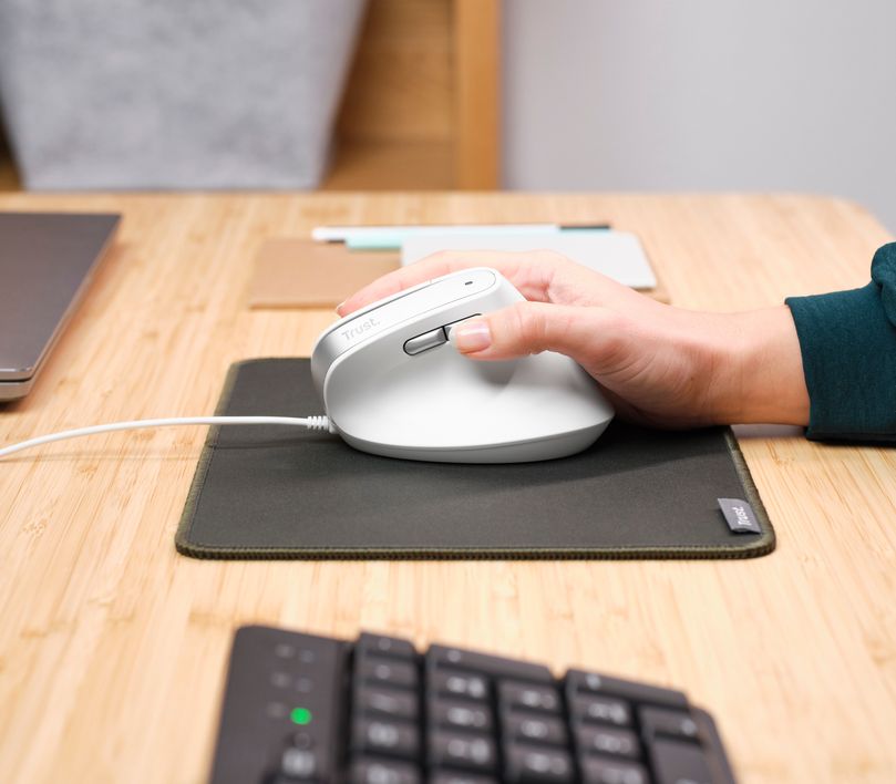 Una mano usando un Mouse ergonómico Trust Bayo II - blanco sobre una alfombrilla oscura en un escritorio de madera. Se puede ver un teclado desenfocado.