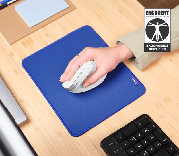 Ratón inalámbrico ergonómico Trust Bayo II blanco sostenido por una mano sobre un mousepad azul en un escritorio de madera. Arriba a la derecha, el sello de certificacion de ERGOCERT. El ratón es de diseño vertical y la mano lo agarra cómodamente. Se ve parte de un teclado negro a la derecha.