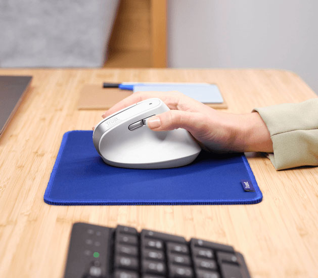 Ratón inalámbrico ergonómico Trust Bayo II blanco en uso, sostenido por una mano, sobre un mousepad azul en un escritorio de madera. El ratón es de diseño vertical y la mano lo agarra cómodamente. Se ve parte de un teclado negro en primer plano y otros objetos desenfocados en el fondo.