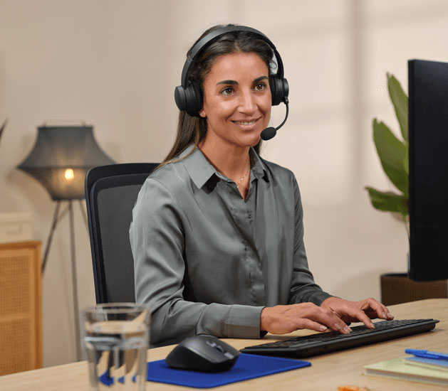 Mujer usando auriculares Trust con micrófono mientras trabaja frente a una computadora en un entorno de oficina hogareña.