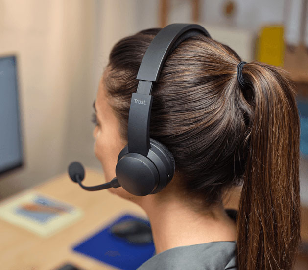 Vista trasera de una mujer usando auriculares Trust con micrófono en una oficina, mostrando el diseño del dispositivo.