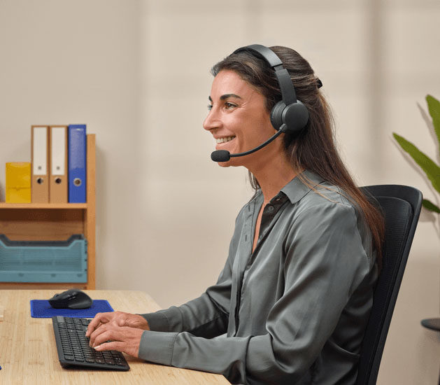 Mujer sonriente usando auriculares Trust con micrófono, escribiendo en teclado en una oficina organizada.