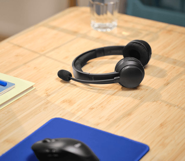 Auriculares Trust con micrófono descansando sobre un escritorio de madera junto a un mouse y un vaso de agua.