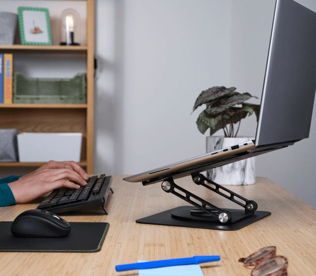 Persona trabajando en una notebook colocada sobre el soporte elevador metálico Zeff de Trust, en un escritorio de madera con teclado y mouse, dentro de un entorno ordenado y moderno.