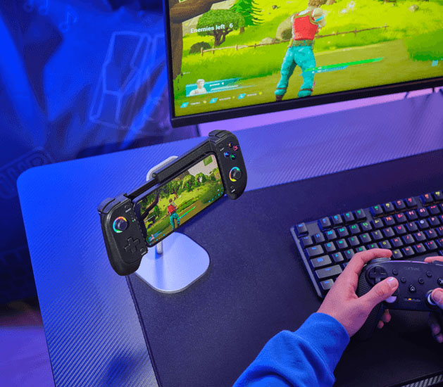 Controlador de juegos móvil Trust GXT 735 Mylox inalámbrico en un soporte de escritorio, con un teléfono inteligente en el medio que muestra un juego. A la derecha, se ve una mano sosteniendo otro control más grande, y en el fondo, un monitor de computadora con el mismo juego. La escena está iluminada en azul y púrpura.