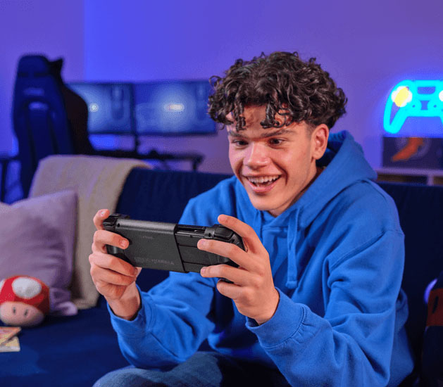 Un joven de pelo rizado, sonriendo y con un buzo azul, jugando con el controlador móvil inalámbrico Trust GXT 735 Mylox acoplado a un teléfono. Está sentado en un sillón oscuro con la luz azul de un monitor y luces de neón en forma de control de fondo.
