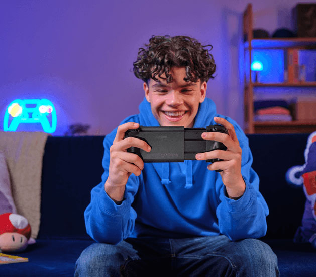 Una toma en primer plano desde atrás de un joven jugando con el controlador móvil inalámbrico Trust GXT 735 Mylox. El controlador sostiene un teléfono en el medio que muestra una escena de juego colorida y el joven viste un buzo azul y jeans.