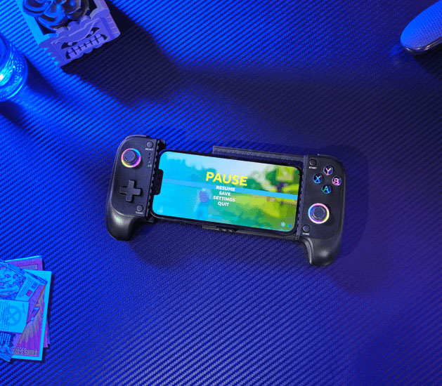 El controlador móvil inalámbrico Trust GXT 735 Mylox en primer plano, sujetando un teléfono que muestra el menú de pausa de un juego. El controlador tiene iluminación RGB en los joysticks y botones, y está sobre una alfombrilla de escritorio azul con textura.