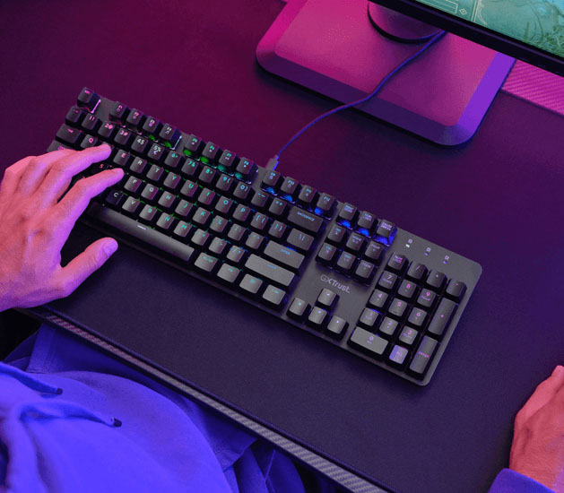 Persona usando un Teclado Mecánico Trust GXT 871 Zora de tamaño completo, conectado por cable a una PC, iluminado con luces RGB, en un entorno con luz violeta y rosa.