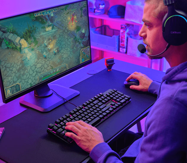 Jugador con auriculares Trust usando un Teclado Mecánico Trust GXT 871 Zora y mouse mientras juega en una PC, en un escritorio con iluminación violeta y rosa, mostrando un videojuego tipo MOBA en pantalla.