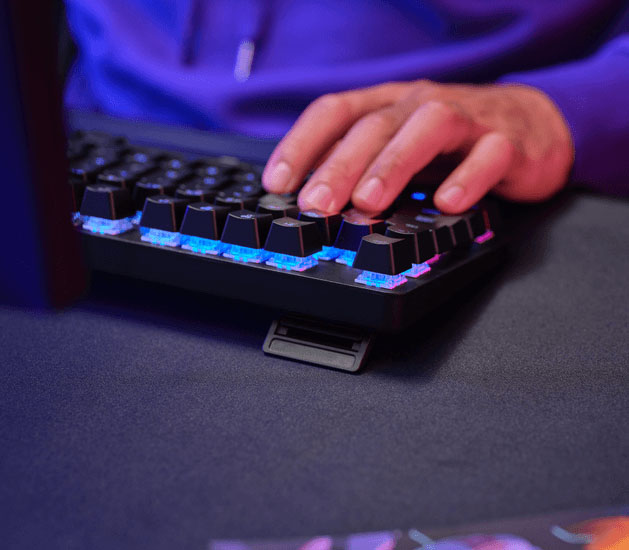 Primer plano de una mano sobre un Teclado Mecánico Trust GXT 871 Zora con retroiluminación RGB azul y rosa y la patita de inclinación desplegada