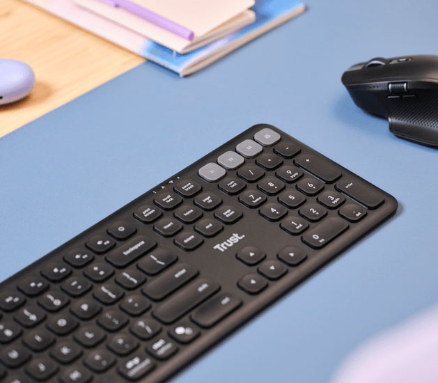 Teclado inalámbrico multidispositivo Trust Vaiya sobre un escritorio celeste, junto a un mouse negro y algunos cuadernos apilados.