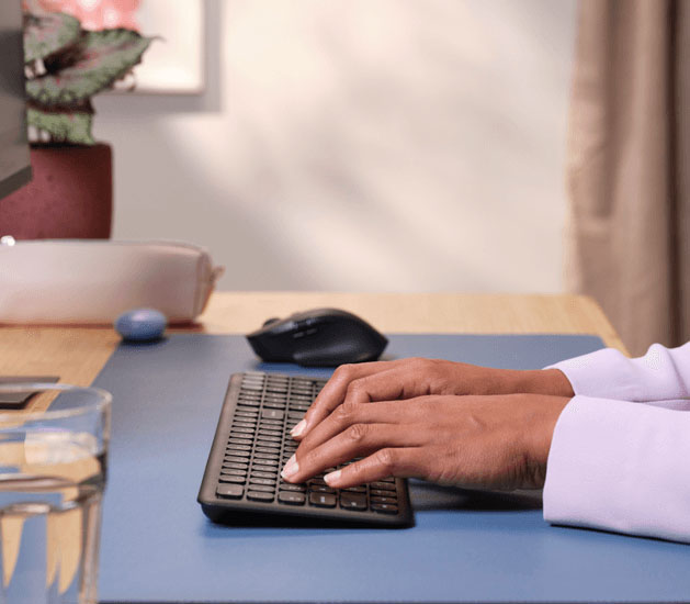 Persona escribiendo en un Teclado inalámbrico multidispositivo Trust Vaiya, sobre un escritorio ordenado con alfombrilla celeste, en un ambiente iluminado y minimalista.