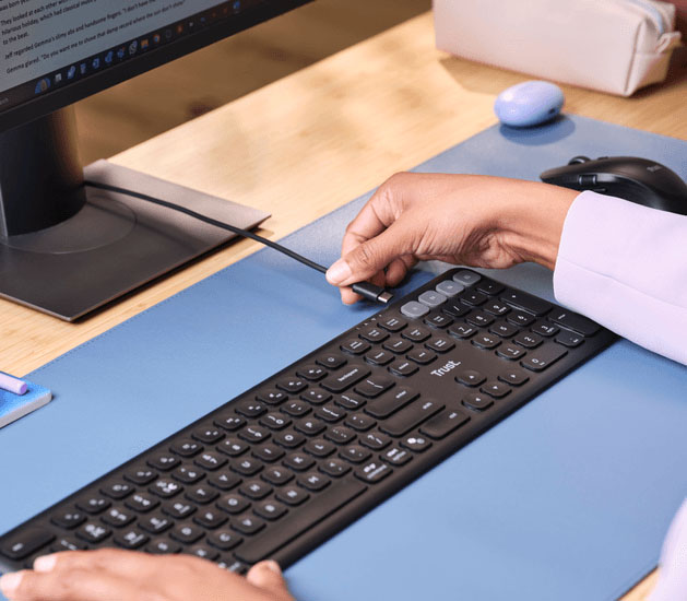 Persona conectando un cable al Teclado inalámbrico multidispositivo Trust Vaiya sobre un escritorio ordenado, con alfombrilla celeste y monitor encendido al fondo.