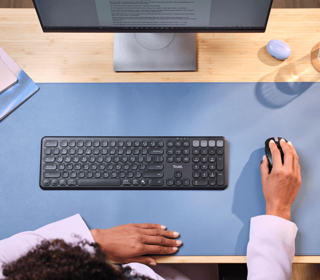 Vista superior de una persona usando un mouse junto a un Teclado inalámbrico multidispositivo Trust Vaiya sobre un escritorio de madera con alfombrilla celeste y monitor encendido.
