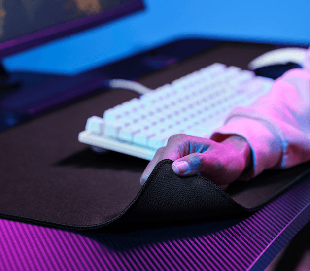 Mano levantando una esquina del mousepad XXL para mostrar la base de goma antideslizante, con un teclado al fondo