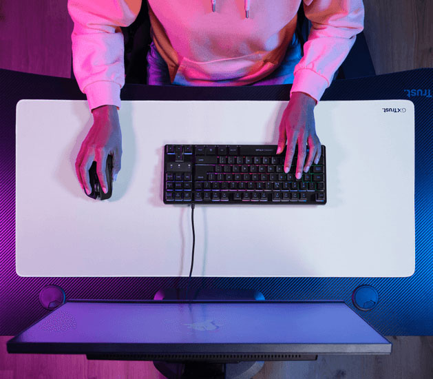 Vista superior de un escritorio con un mousepad XXL blanco, teclado y mouse, con una persona apoyando las manos sobre los periféricos.