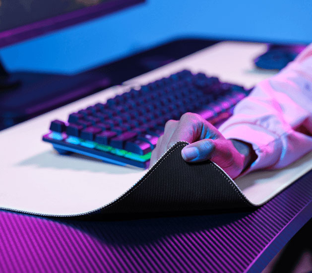 Mano levantando una esquina del mousepad XXL para mostrar la base de goma antideslizante, con un teclado al fondo