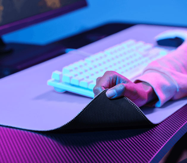 Mano levantando una esquina del mousepad XXL para mostrar la base de goma antideslizante, con un teclado al fondo