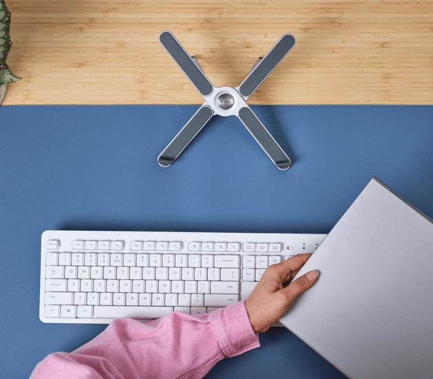 Vista superior del soporte plegable de aluminio Macy de Trust en forma de X sobre un escritorio, con una mano acercando una notebook y un teclado blanco en primer plano.