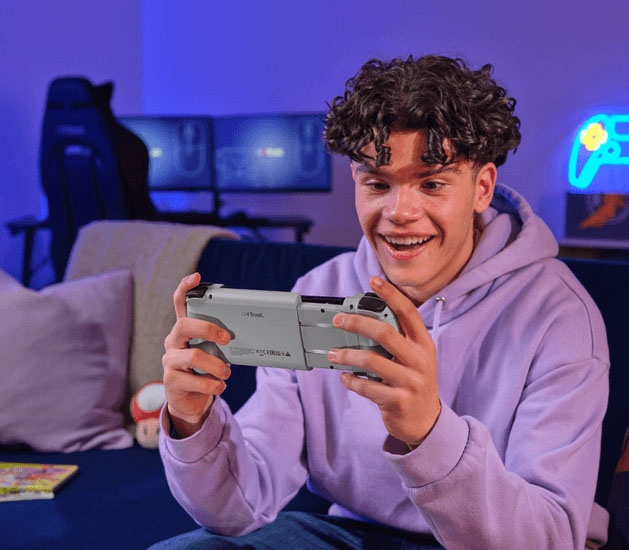 Un joven sonriente con una sudadera con capucha lila está sentado en un sofá oscuro y juega con el controlador Trust GXT 735G Mylox inalámbrico para juegos móviles (gris), que tiene un teléfono móvil acoplado en el centro. El joven está enfocado en la pantalla y tiene una expresión de disfrute. En el fondo, hay una silla gamer, dos monitores de PC apagados o con poca luz, y un cartel de neón con la forma de un controlador de juego. El ambiente está iluminado con luces azules y moradas.