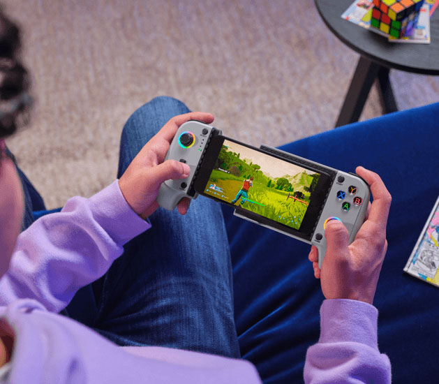 Manos de un joven sosteniendo el controlador Trust GXT 735G Mylox inalámbrico para juegos móviles (gris) que tiene un teléfono móvil acoplado en el centro, mostrando un juego de estilo cartoon en pantalla, con un personaje en un entorno exterior verde. El jugador está sentado y usa jeans azules y una sudadera con capucha lila, con un cubo de Rubik y otros objetos fuera de foco en el fondo.