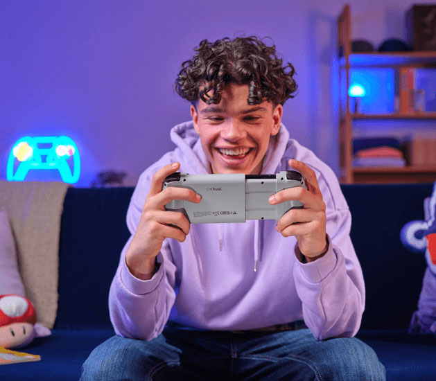 Primer plano de un joven sonriente, concentrado en jugar con el controlador Trust GXT 735G Mylox inalámbrico para juegos móviles (gris). El joven está sentado en un sofá oscuro y usa una sudadera con capucha lila. Se puede ver la parte posterior del controlador con la marca GXT Trust y las certificaciones CE/FCC, junto con un neón azul de un controlador en el fondo.