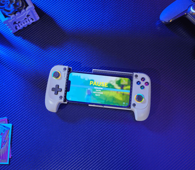 Primer plano cenital del controlador Trust GXT 735G Mylox inalámbrico para juegos móviles (gris) con un teléfono móvil acoplado en el centro, mostrando una pantalla de pausa con las opciones reanudar, guardar, ajustes y salir. La superficie es un escritorio azul oscuro con un patrón de fibra de carbono y hay otros accesorios de juego fuera de foco, incluyendo cartas coleccionables y la parte superior de otro controlador. Los joysticks del controlador tienen un sutil anillo de luz RGB.