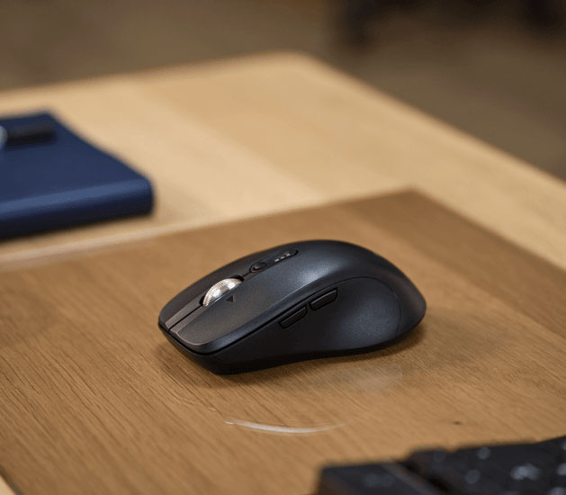 Mouse inalámbrico Trust Ferro Hyperscroll negro sobre un escritorio de madera con una alfombrilla de ratón marrón claro. El mouse tiene un diseño ergonómico y varios botones, incluyendo una rueda de desplazamiento plateada.