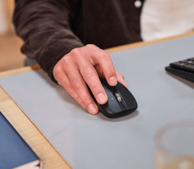 Primer plano de una persona usando el mouse inalámbrico Trust Ferro Hyperscroll negro sobre una alfombrilla de ratón gris. Se ve la mano de la persona sujetando el mouse y el pulgar apoyado en el lateral.