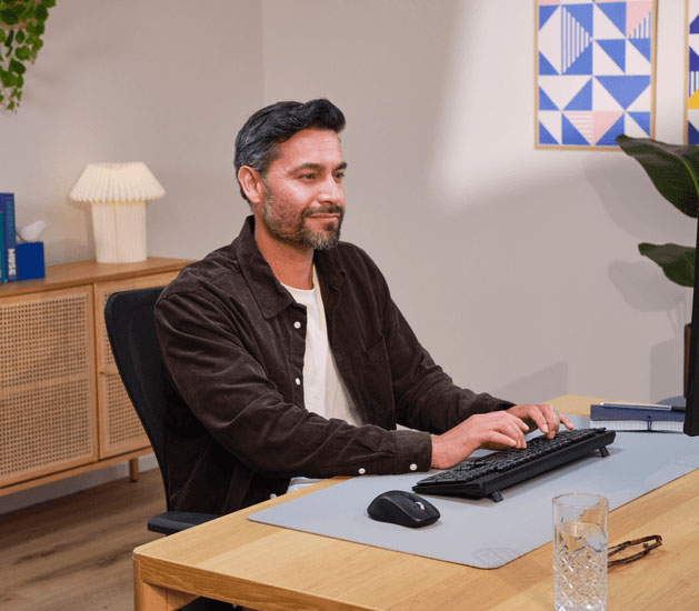 Hombre sonriente usando la computadora en un escritorio de madera, tecleando en un teclado negro con el mouse inalámbrico Trust Ferro Hyperscroll al lado, sobre una alfombrilla gris.