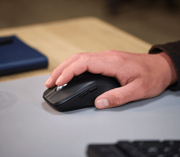 Primer plano de una persona usando el mouse inalámbrico Trust Ferro Hyperscroll negro sobre una alfombrilla de ratón gris. Se ve la mano de la persona y el dedo índice tocando la rueda de desplazamiento plateada del mouse.