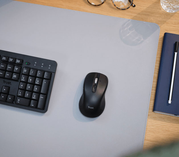 Primer plano del mouse inalámbrico Trust Ferro Hyperscroll negro, un teclado Trust negro, un cuaderno azul, un bolígrafo y anteojos sobre una alfombrilla de ratón gris en un escritorio de madera.