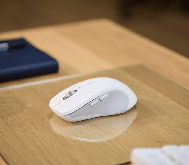 Mouse inalámbrico Trust Ferro Hyperscroll blanco sobre un escritorio de madera con un mousepad transparente. Se ve un objeto azul desenfocado en la parte superior izquierda.