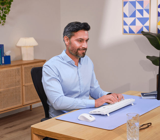Mouse inalámbrico Trust Ferro Hyperscroll blanco sobre un escritorio de madera con un mousepad transparente. Se ve un objeto azul desenfocado en la parte superior izquierda y parte de un teclado blanco desenfocado en la inferior derecha.