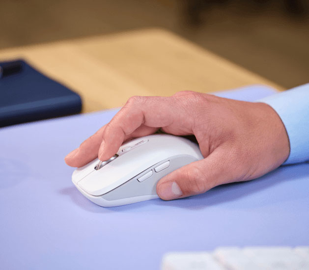 Hombre sonriente usando la computadora en un escritorio de madera, tecleando en un teclado blanco con el mouse inalámbrico Trust Ferro Hyperscroll blanco al lado, sobre un mousepad lila.