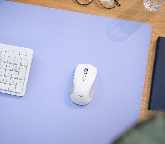 Vista aérea del mouse inalámbrico Trust Ferro Hyperscroll blanco y un teclado blanco a la izquierda sobre un mousepad lila. Se ven anteojos, un cuaderno y un vaso de agua en el escritorio de madera.