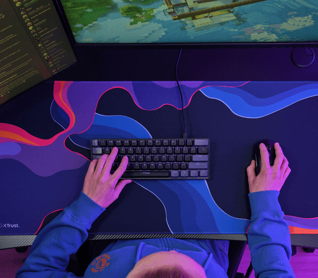 Vista superior de un escritorio con mousepad Trust GXT 759 XXL azul y violeta, teclado y mouse, con una persona usando ambos periféricos.