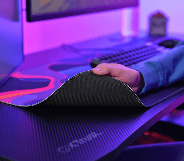 Mano levantando una esquina del mousepad Trust GXT 759 XXL para mostrar la base de goma antideslizante, con teclado al fondo.