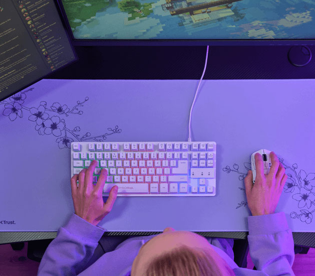 Vista superior de un escritorio con mousepad Trust GXT 759 XXL violeta y magenta, teclado y mouse, con una persona usando ambos periféricos.