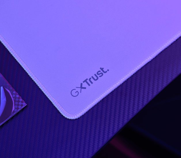 Detalle del mousepad Trust GXT en violeta y magenta con bordes cosidos y logo GXT Trust en la esquina.