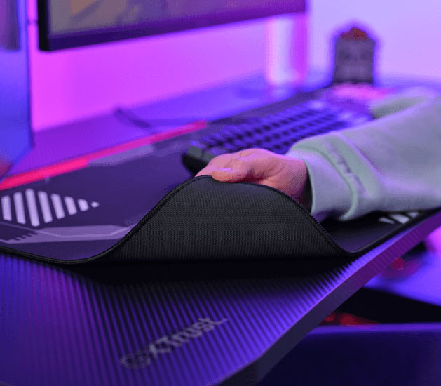 Mano levantando una esquina del mousepad Trust GXT 759 XXL para mostrar la base de goma antideslizante, con teclado al fondo.