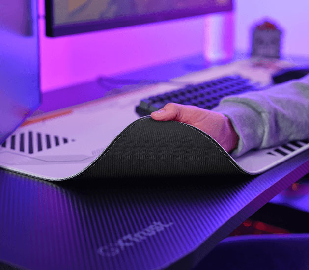 Mano levantando una esquina del mousepad Trust GXT 759 XXL para mostrar la base de goma antideslizante, con teclado al fondo.