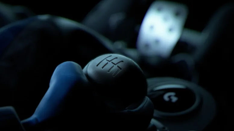 Primerísimo plano de la palanca de cambios Logitech Driving Force™ Shifter en la oscuridad, con el patrón de seis velocidades en la parte superior y un pedal iluminado al fondo