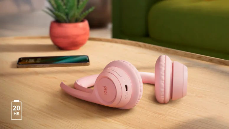 Auriculares inalámbricos Logitech Zone 300 de color rosa pastel plegados sobre una mesa de madera junto a un smartphone y una maceta con una planta. En la esquina inferior izquierda, un ícono de batería indica 20 horas de autonomía