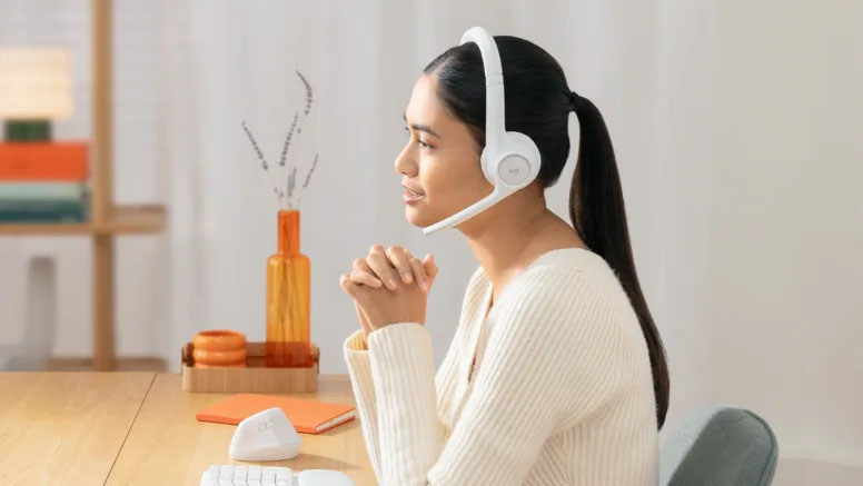 Mujer joven de perfil, sentada en un escritorio en un entorno hogareño, usando los auriculares inalámbricos Logitech Zone 300 blancos con el micrófono bajado, mirando hacia el frente con las manos entrelazadas sobre el escritorio.