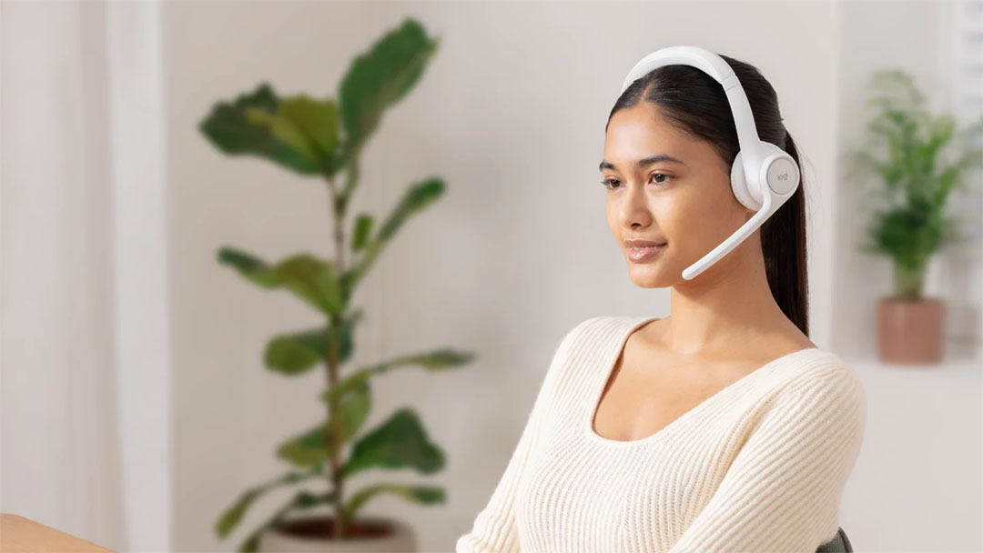 Mujer joven sentada en un interior luminoso con una planta grande detrás, usando los auriculares inalámbricos Logitech Zone 300 blancos con el micrófono bajado, sonriendo levemente.